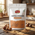 A stand-up pouch of Nature's Basket Aritha Powder natural shampoo, featuring a white and orange label with product details and illustrations of aritha seeds, set against a solid beige background.